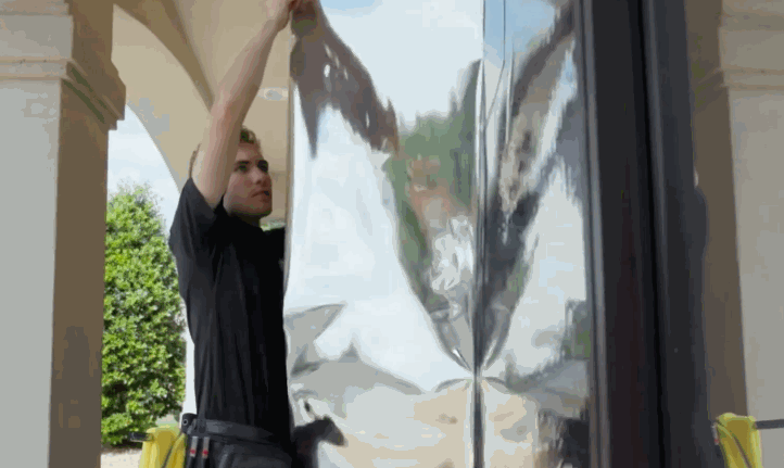 A technician carefully aligning and installing reflective window film on a tall window, highlighting precision and expertise