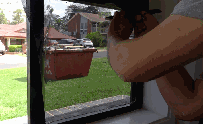 A person carefully applies tinted film to a small residential window, with a suburban neighborhood visible outside